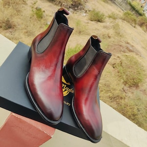 May include: Burgundy leather Chelsea boots with elastic side panels. The boots have a polished finish and are displayed on a black box. The boots have a dark brown sole and a pull tab at the back.