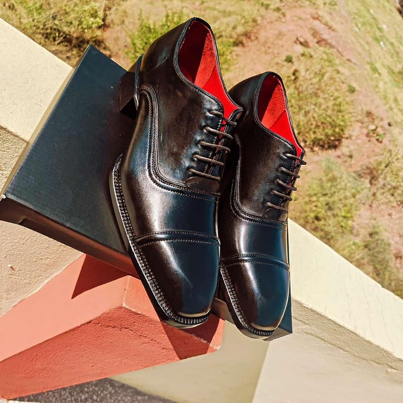 May include: A pair of black leather dress shoes with a high-gloss finish. The shoes have a classic Oxford design with a cap toe, lace-up closure, and a red interior. The shoes are displayed on a black box.