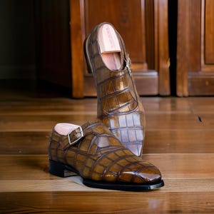 May include: A pair of brown leather dress shoes with a crocodile pattern. The shoes have a buckle closure and a wooden shoe tree inside. The shoes are displayed on a wooden floor.