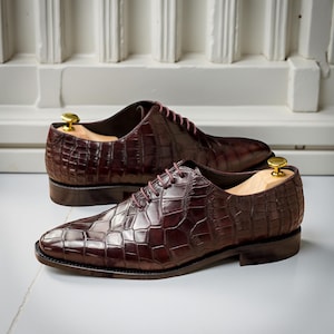 Puede incluir: Un par de zapatos de vestir de cuero marr&oacute;n con un patr&oacute;n texturizado similar al de un cocodrilo. Los zapatos tienen un dise&ntilde;o Oxford cl&aacute;sico con cordones y se exhiben con hormas de madera en su interior. El fondo es una pared blanca.