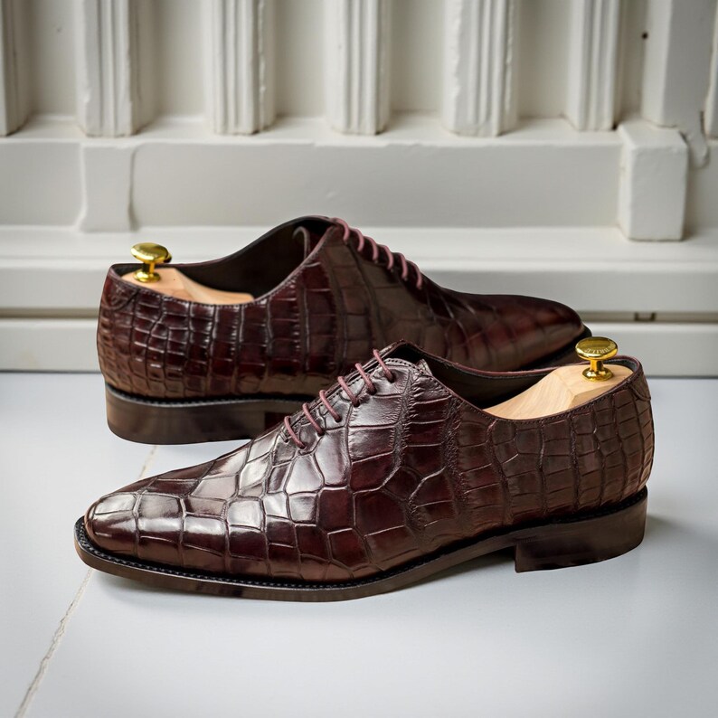 Puede incluir: Un par de zapatos de vestir de cuero marr&oacute;n con un patr&oacute;n texturizado similar al de un cocodrilo. Los zapatos tienen un dise&ntilde;o Oxford cl&aacute;sico con cordones y se exhiben con hormas de madera en su interior. El fondo es una pared blanca.