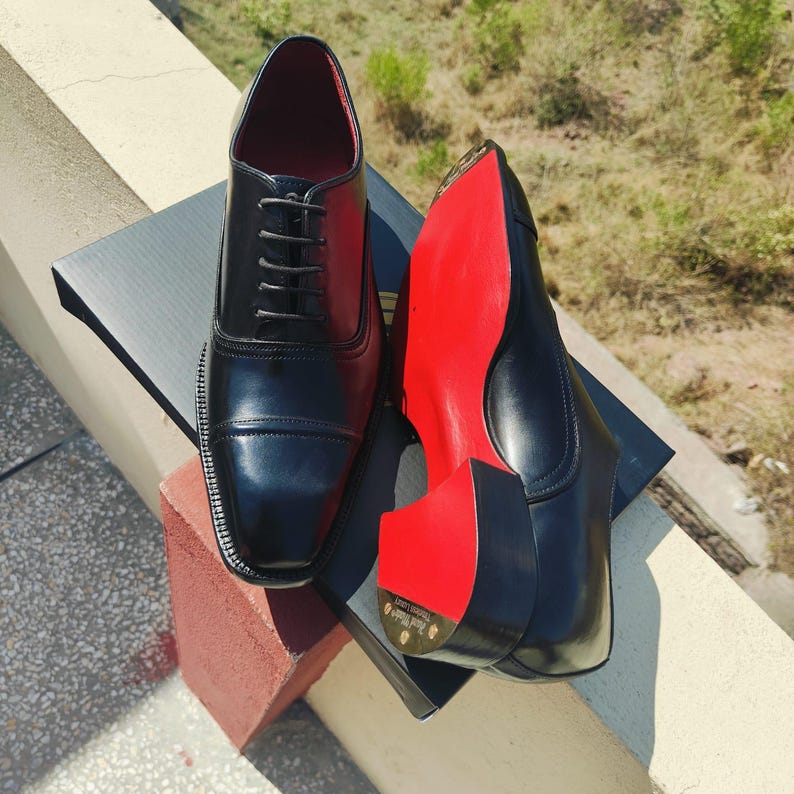May include: Black leather dress shoes with red soles. The shoes have a classic Oxford design with a cap toe and lace-up closure. The interior is red. The shoes are displayed on a black box.