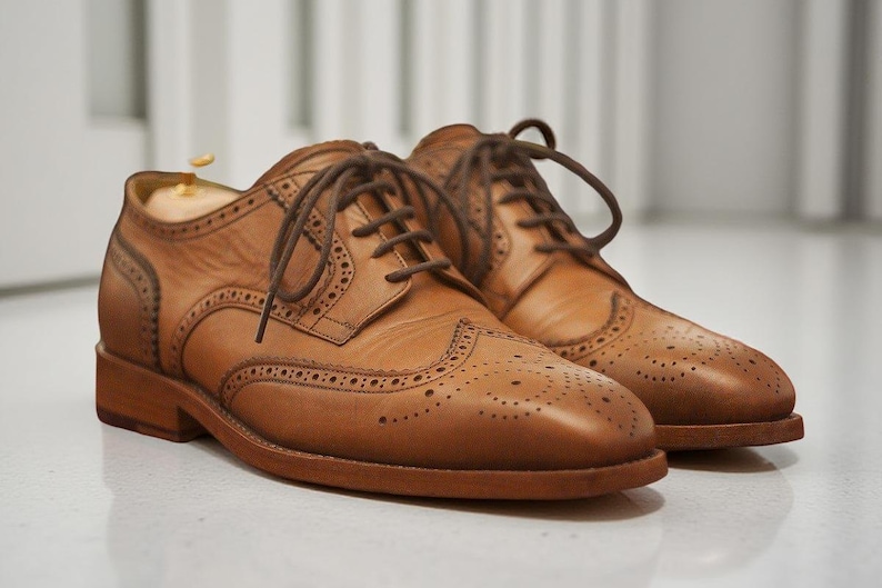 K&ouml;nnte beinhalten: Ein Paar hellbraune Leder-Brogue-Schuhe mit braunen Schn&uuml;rsenkeln. Die Schuhe haben dekorative Perforationen und stehen auf einer hellen Oberfl&auml;che. Klassischer Stil.