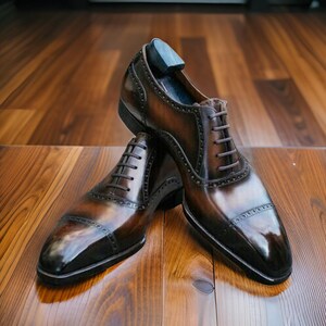 Op de afbeelding: Een paar bruine leren herenschoenen met een glanzende afwerking. De schoenen hebben een klassiek Oxford-ontwerp met ingewikkelde stiksels en een neuskap. De veters zijn bruin en de schoenen staan op een houten oppervlak.