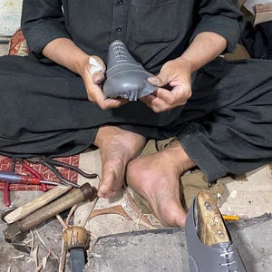 May include: A shoemaker at work, seated on the floor, holding a gray shoe last. Various tools, including hammers, pliers, and knives, are scattered around. A partially constructed shoe is visible, along with other shoe components.