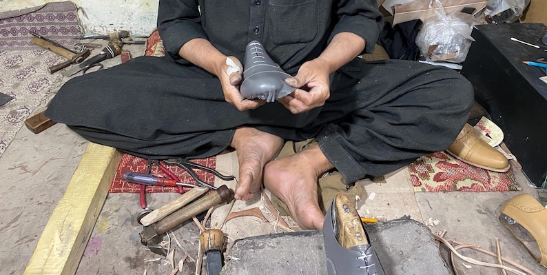 May include: A shoemaker at work, seated on the floor, holding a gray shoe last. Various tools, including hammers, pliers, and knives, are scattered around. A partially constructed shoe is visible, along with other shoe components.