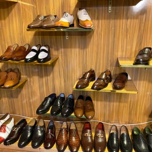 May include: A display of various men's dress shoes on wooden shelves. Styles include loafers, oxfords, and monk straps in shades of brown, black, and white. Some shoes have decorative details like buckles and stitching.