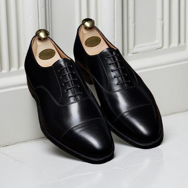 May include: A pair of black leather dress shoes with a cap toe design. The shoes have a classic lace-up closure and are polished to a high shine. Shoe trees are inserted inside the shoes. The shoes are displayed on a white surface.