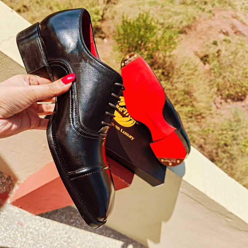 May include: A pair of black leather dress shoes with a glossy finish. One shoe is held up, revealing a red interior and a brown sole. The other shoe is in the background, showing a red sole with the text "Dress Luxury".