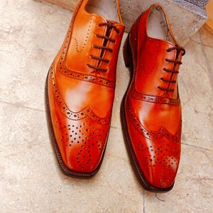 May include: A pair of orange leather dress shoes with intricate brogue detailing. The shoes feature a classic wingtip design with decorative perforations and dark brown laces. The polished leather reflects light, highlighting the shoe's craftsmanship.