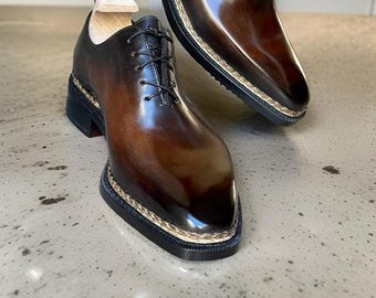 Op de afbeelding: Een paar bruine leren herenschoenen met een vierkante neus en zwarte veters. De schoenen hebben een dikke zool met een decoratieve gestikte rand. Het leer heeft een gepolijste, glanzende afwerking.