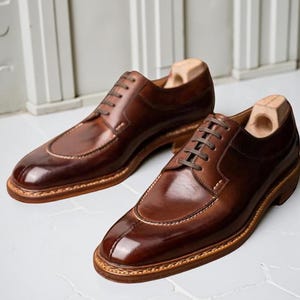Peut inclure: Une paire de chaussures de ville en cuir marron brillant. Les chaussures présentent un design classique avec un bout arrondi, une fermeture à lacets et une semelle cousue. Des embauchoirs sont insérés dans les chaussures. Les chaussures sont présentées sur une surface blanche.