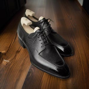 Op de afbeelding: Een paar zwarte leren herenschoenen met een getextureerde afwerking en een glanzende neus. De schoenen zijn geregen en hebben houten schoenspanners. Ze staan op een houten oppervlak.