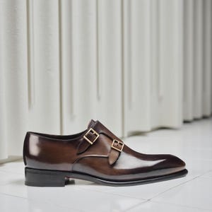 K&ouml;nnte beinhalten: Ein brauner Lederschuh mit doppeltem Monk-Strap-Verschluss. Der Schuh hat eine spitze Zehenpartie und goldfarbene Schnallen. Das Leder hat eine polierte, gl&auml;nzende Oberfl&auml;che, und der Schuh steht auf einem niedrigen, schwarzen Absatz.