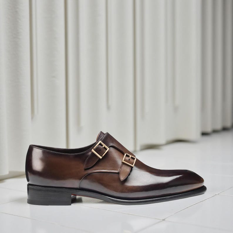 K&ouml;nnte beinhalten: Ein brauner Lederschuh mit doppeltem Monk-Strap-Verschluss. Der Schuh hat eine spitze Zehenpartie und goldfarbene Schnallen. Das Leder hat eine polierte, gl&auml;nzende Oberfl&auml;che, und der Schuh steht auf einem niedrigen, schwarzen Absatz.