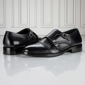 May include: A pair of black leather double monk strap shoes. The shoes feature a cap toe design, decorative perforations, and silver-tone buckles. The shoes are displayed on a reflective surface.