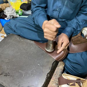 May include: A person seated, working on a brown leather shoe with a metal tool. The shoe is in the process of being made, with other shoe components and tools visible. The person is wearing a blue outfit.