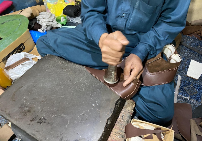 May include: A person seated, working on a brown leather shoe with a metal tool. The shoe is in the process of being made, with other shoe components and tools visible. The person is wearing a blue outfit.