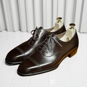 May include: A pair of brown leather Oxford shoes with a cap toe design and lace-up closure. The shoes are displayed with wooden shoe trees inside, showcasing their shape and structure. The polished leather reflects light, highlighting the details.