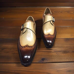 May include: A pair of two-tone leather dress shoes with a monk strap buckle closure. The shoes feature a light tan upper with a dark brown toe and decorative perforations along the edges. The shoes are displayed on a wooden surface.