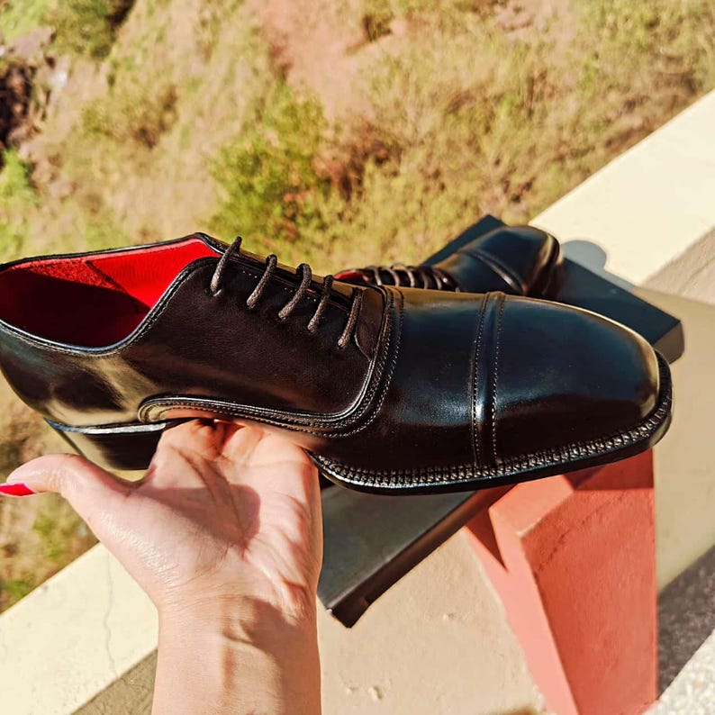 May include: A pair of black leather dress shoes with a cap toe design. The shoes have a glossy finish, a red interior, and brown laces. The shoes are displayed on a black surface.