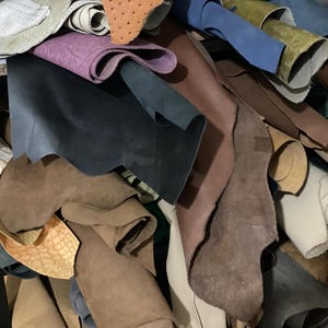 May include: A pile of various leather hides in shades of brown, blue, purple, and tan. Some hides are rolled, while others are flat, showcasing different textures and finishes. The image suggests a craft supply or material for leatherworking.