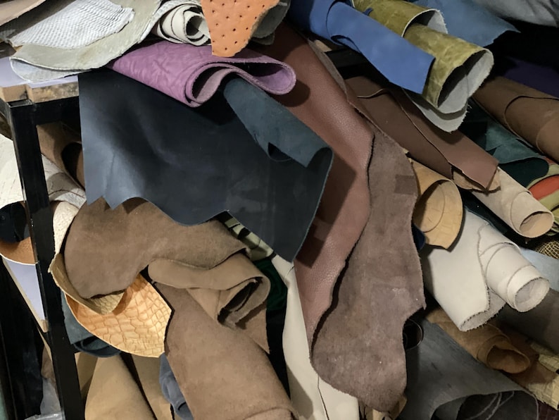 May include: A pile of various leather hides in shades of brown, blue, purple, and tan. Some hides are rolled, while others are flat, showcasing different textures and finishes. The image suggests a craft supply or material for leatherworking.