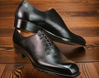May include: Two black leather dress shoes are stacked on a wooden surface. The top shoe is a slip-on style, while the bottom shoe has laces. Both shoes have a glossy finish and a low heel. The background is a warm, brown wood.