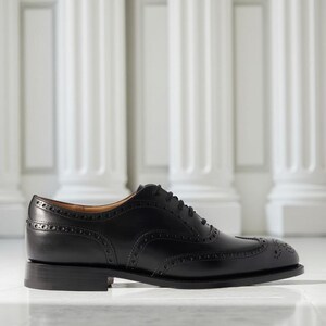 May include: A black leather Oxford shoe with brogue detailing. The shoe features a classic wingtip design and a low heel. The shoe is displayed on a reflective surface with white columns in the background.