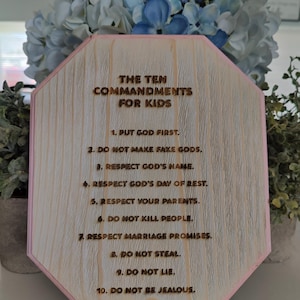 May include: An octagonal wooden plaque with a light pink border, featuring the laser-engraved text THE TEN COMMANDMENTS FOR KIDS followed by a numbered list from 1 to 10, resting on a small white plastic stand.