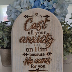 May include: Wooden plaque with the text "Cast all your anxiety on Him because He cares for you." in a decorative font. The plaque is supported by a white stand and has a light wood grain finish. The text is in a darker brown colour.