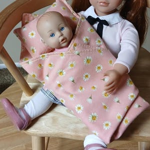 May include: A doll with red hair and blue eyes sits on a wooden chair, holding a smaller doll wrapped in a pink blanket with white daisy patterns. The dolls are wearing pink shoes and white socks.