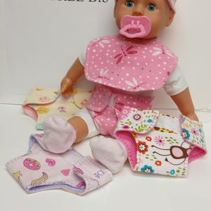 May include: A doll wearing a pink hat with a bow, a pink pacifier, and a pink dragonfly-patterned bib. The doll is dressed in a white shirt and a pink patterned diaper. Two additional diapers with colorful prints are placed in front of the doll.