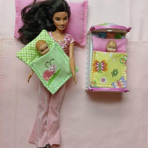 May include: A doll with long black hair wearing a pink and white top and pink pants, holding a green pillow with a ladybug design. A baby doll is nestled in the pillow. A miniature bed with a baby doll inside is also present.