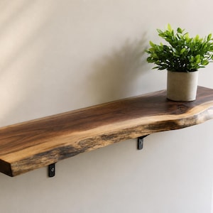 May include: A live edge wooden wall shelf with a dark brown grain and natural bark texture on the front edge. The shelf is mounted to the wall with two small black metal brackets and holds a small green potted plant in a light grey container.