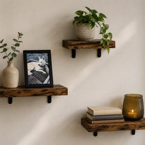 May include: Three rectangular wall-mounted shelves made of dark brown wood with visible grain and black metal brackets. The shelves hold a beige vase with greenery, a black picture frame, a potted plant, a stack of three books, and a ribbed amber glass candle holder.