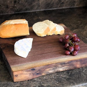 May include: A wooden serving board with bread, sliced bread, cheese, and red grapes. The board is rectangular with a natural edge and a dark brown finish. The bread is a golden brown colour.