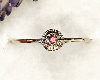 May include: A delicate gold ring with a textured, circular setting featuring a small, red gemstone.