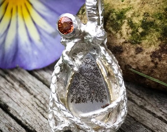 Puede incluir: Un colgante de plata con una piedra de ágata dendrítica engastada en una jaula de alambre de plata y oro. El colgante tiene una pequeña piedra preciosa naranja en la parte superior.