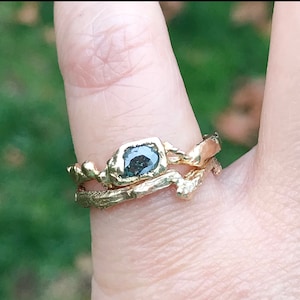 May include: A gold ring with a dark gray, faceted gemstone set in a rough, organic-shaped band. The ring is worn on a finger with a second, thinner gold band.
