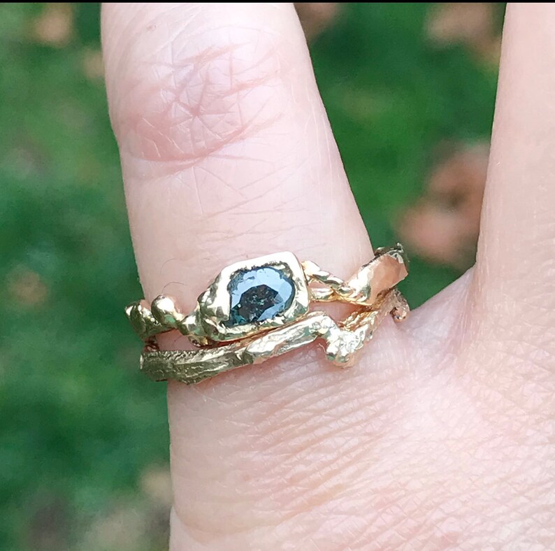 May include: A gold ring with a dark gray, faceted gemstone set in a rough, organic-shaped band. The ring is worn on a finger with a second, thinner gold band.