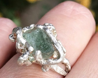 May include: A silver ring with a rough, organic design. The ring features a rectangular, pale green gemstone set within the textured metal. The band has a unique, irregular shape, adding to the ring's handcrafted aesthetic.