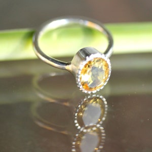 Puede incluir: Un anillo de plata con una piedra preciosa amarilla engastada en un engaste de bisel. El anillo est&aacute; sobre una superficie verde.