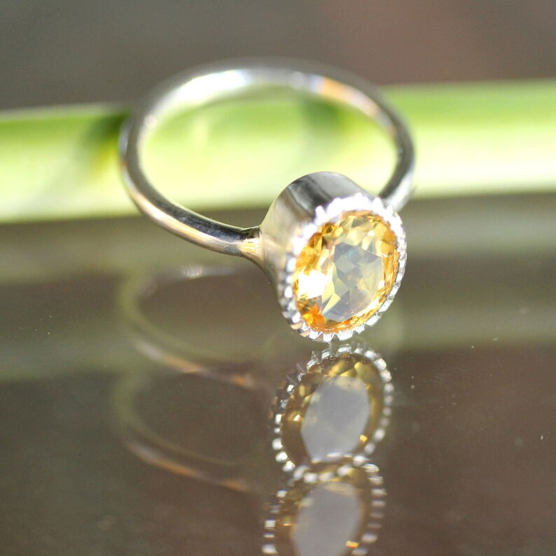 Puede incluir: Un anillo de plata con una piedra preciosa amarilla engastada en un engaste de bisel. El anillo est&aacute; sobre una superficie verde.