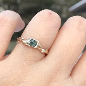 May include: A gold ring with a unique, organic design features a dark green gemstone set in a rough, textured bezel. The ring is worn on a person's finger.