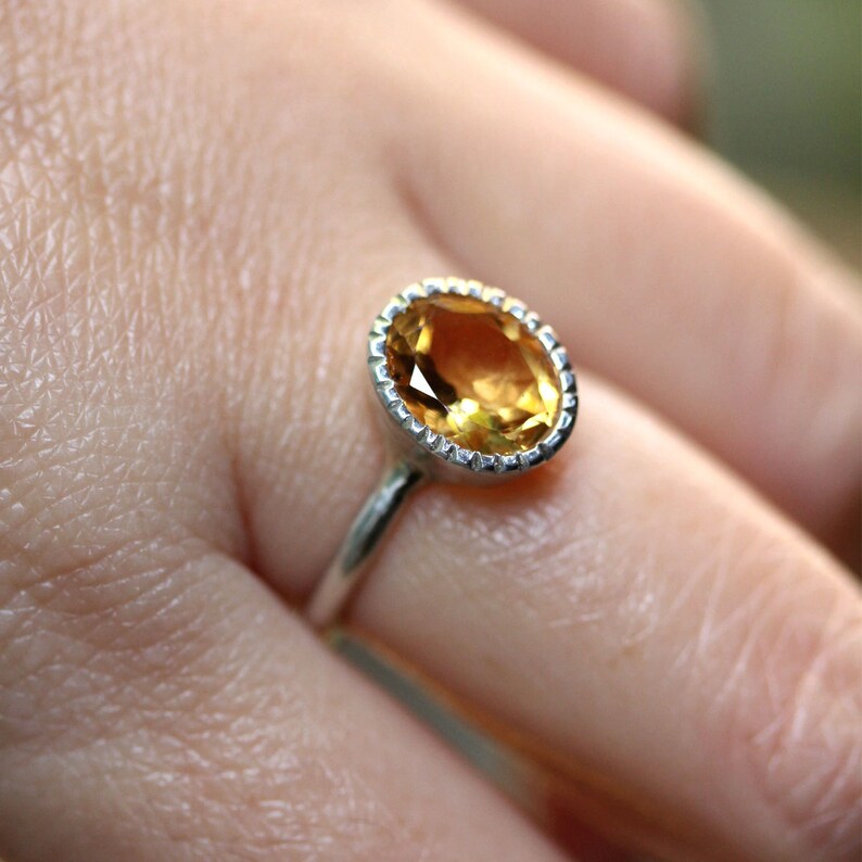 Puede incluir: Un anillo de plata con una gran piedra preciosa amarilla de forma ovalada engastada en un engaste de bisel. El anillo est&aacute; en un dedo.