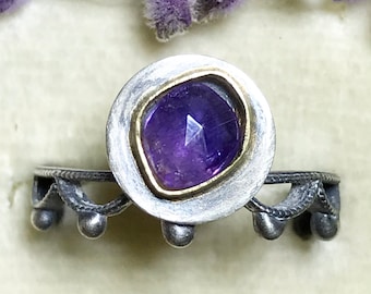 May include: A silver ring with a purple amethyst gemstone set in a gold bezel. The ring has a delicate, ornate band with small silver beads.