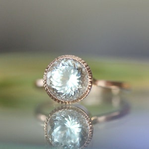 May include: A delicate ring with a round, clear aquamarine gemstone set in a rose gold bezel setting. The ring has a textured band.