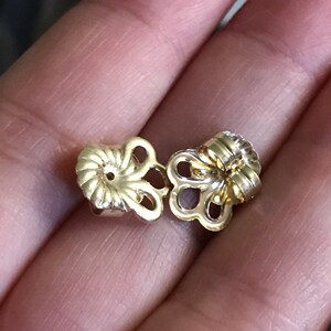 May include: A pair of gold-tone earring backs with a decorative design. The backs are designed to secure earrings in place.