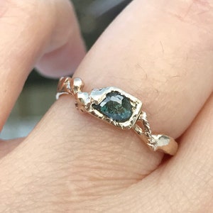 May include: A gold ring with a green gemstone set in a textured band. The ring is worn on a finger.